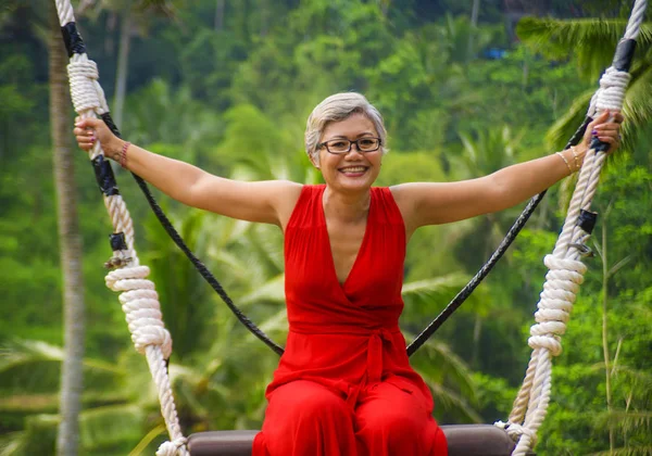 Retrato estilo de vida natural de atraente feliz meia idade 40s - 50s mulher asiática com cabelos grisalhos e elegante vestido vermelho equitação floresta tropical swing despreocupado balançando — Fotografia de Stock