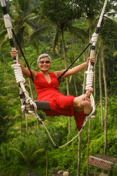 Retrato estilo de vida natural de atraente feliz meia idade 40s - 50s mulher asiática com cabelos grisalhos e elegante vestido vermelho equitação floresta tropical swing despreocupado balançando — Fotografia de Stock