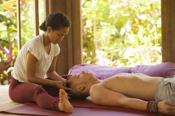 Junge schöne und exotische asiatische thailändische Therapeutin, die dem kaukasischen Mann im Heilbad für alternative Medizin traditionelle Kopf- und Gesichtsmassage gibt — Stockfoto