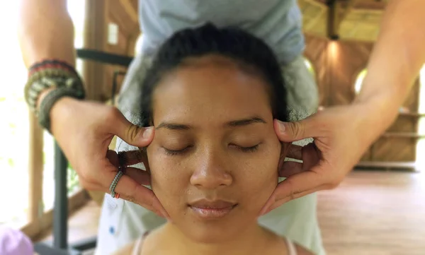 Primer plano retrato de la cara de joven hermosa y relajada mujer indonesia asiática que recibe masaje tailandés facial tradicional con manos masculinas trabajando su cabeza —  Fotos de Stock