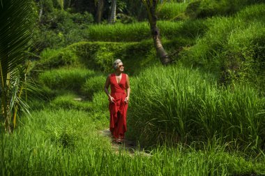 yeşil tropikal orman manzara üzerinde yürüyen şık kırmızı elbise gri saçlı güzel ve mutlu orta yaşlı 40'lı veya 50'li Asyalı kadının doğal yaşam tarzı portresi