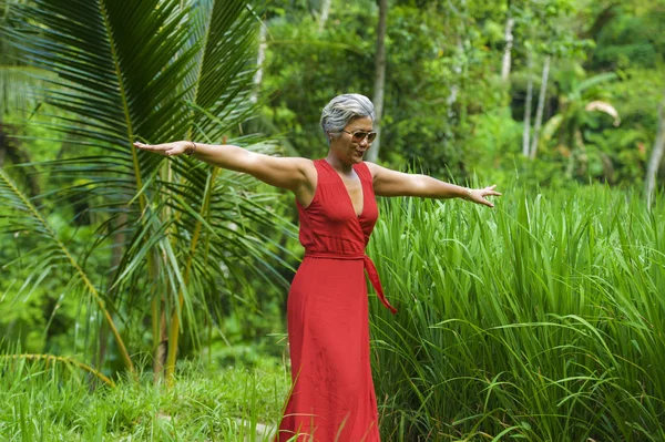 녹색 열대 숲 풍경을 걷는 세련된 빨간 드레스에 회색 머리를 가진 아름답고 행복한 중년 40 대 또는 50 대 아시아 여성의 자연 라이프 스타일 초상화 — 스톡 사진