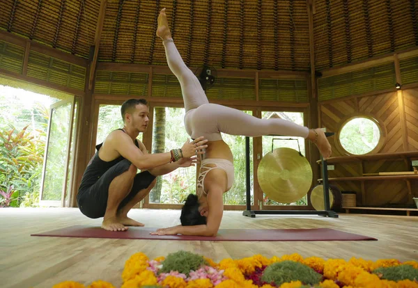 Joven mujer hermosa y en forma con acrobat hombre como entrenador de yoga aprendizaje equilibrio postura en el entrenamiento de acroyoga en el exótico gimnasio en el bienestar y estilo de vida saludable — Foto de Stock