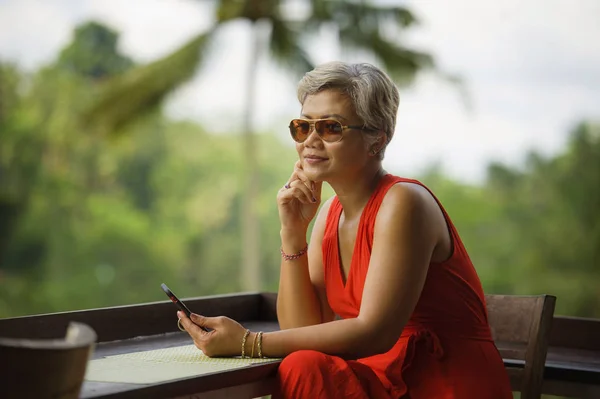 Atractiva y relajada mujer asiática de 40 o 50 años con pelo gris y vestido rojo con estilo utilizando las redes sociales en el teléfono móvil inernet en el hermoso complejo de la selva tropical —  Fotos de Stock