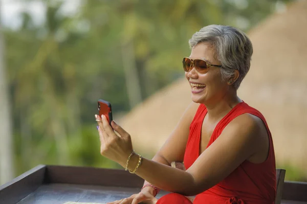 Attractive and relaxed 40s or 50s Asian woman with grey hair and stylish red dress using social media on inernet mobile phone at beautiful tropical jungle resort — Stock Photo, Image