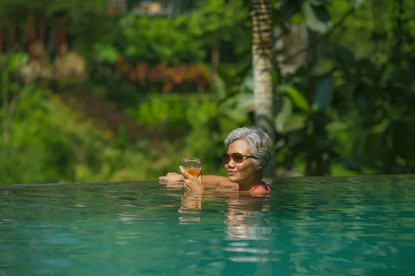 Estilo de vida natural retrato de mujer asiática atractiva y feliz de mediana edad relajada en el complejo tropical infinito simming piscina con fondo de selva disfrutando de una bebida relajada — Foto de Stock