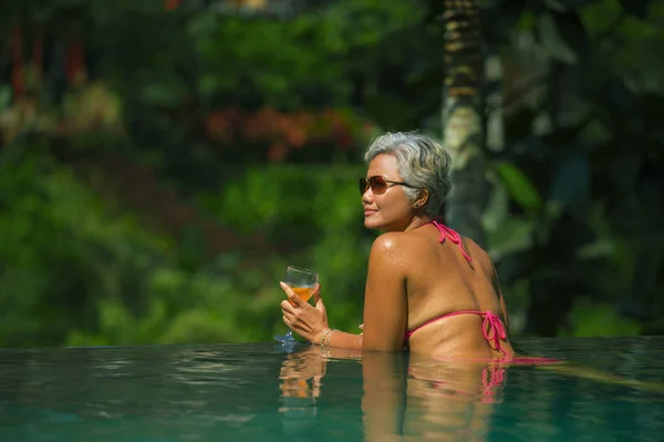 Estilo de vida natural retrato de mujer asiática atractiva y feliz de mediana edad relajada en el complejo tropical infinito simming piscina con fondo de selva disfrutando de una bebida relajada — Foto de Stock