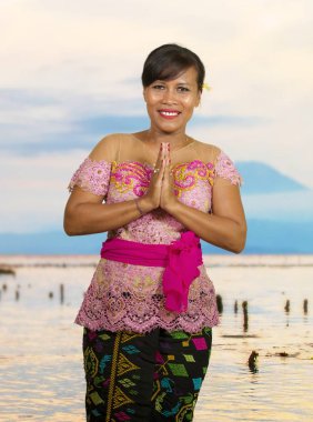 beautiful happy middle aged Indonesian Balinese woman in traditional ceremony dress at tropical beach with Bali Agung volcano background in holidays tourist destination clipart