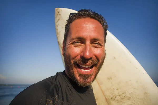 Retrato natural de homem surfista atraente e feliz em seus 40 anos carregando prancha de surf após a manhã de surf na bela praia em wetsuit sorrindo desfrutando de férias de verão — Fotografia de Stock