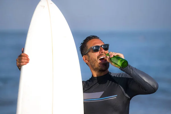 Retrato natural de homem surfista atraente e feliz em seus 40 anos carregando prancha de surf após a manhã de surf na bela praia em roupa de mergulho bebendo garrafa de cerveja desfrutando de verão — Fotografia de Stock