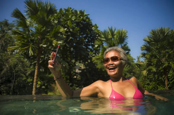 Atractiva y feliz de mediana edad mujer indonesia asiática en bikini nadando en el resort tropical de lujo piscina infinita tomando retrato selfie con teléfono móvil disfrutando de verano — Foto de Stock