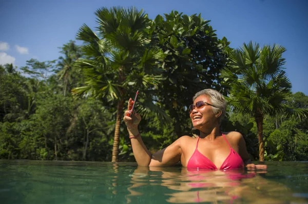 Привлекательная и счастливая азиатская женщина средних лет в бикини купается в тропическом роскошном бассейне бесконечности курорта делая селфи-портрет с мобильным телефоном наслаждаясь летом — стоковое фото