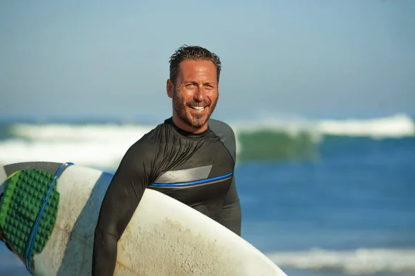 handsome and attractive surfer man in neoprene swimsuit carrying surf board smiling happy and cheerful after surfing enjoying Summer water sport and holidays