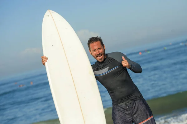 Snygg och attraktiv surfare man i neopren baddräkt innehav surfbräda poserar sval efter surfing njuter av sommar vattensport och helgdagar ger handtecken — Stockfoto