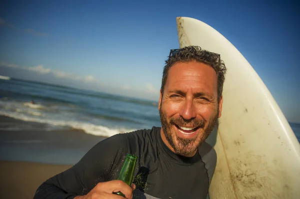 Attraktiva Surfer man i neopren baddräkt Holding Surf Board poserar sval och glad efter surfing njuter sommar vattensport och helgdagar dricka ölflaska — Stockfoto