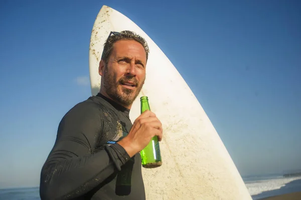 Attraktiva Surfer man i neopren baddräkt Holding Surf Board poserar sval och glad efter surfing njuter sommar vattensport och helgdagar dricka ölflaska — Stockfoto