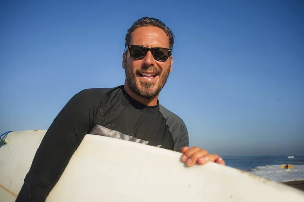 Bonito e atraente surfista homem em neoprene maiô carregando prancha de surf sorrindo feliz e alegre depois de surfar apreciando verão água esporte e feriados — Fotografia de Stock