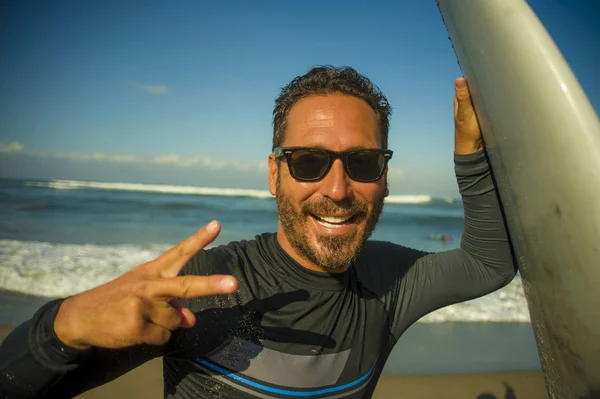 Livsstil porträtt av attraktiva och Happy Surfer man 3os till 40s i neopren surfing baddräkt poserar med surfbräda på stranden njuter vattensport och sommarlov — Stockfoto