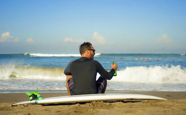 Привабливий і щасливий серфінг чоловік 3os до 40s в неопренових серфінг купальник позує з дошкою для серфінгу, сидячи на пляжному піску насолоджуючись відпочинком пивна пляшка розслаблена — стокове фото