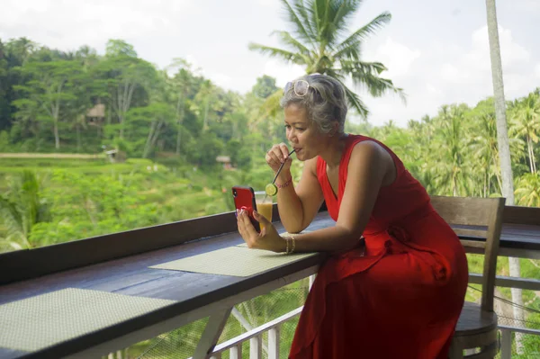 Vonzó és nyugodt 40-es vagy 50-es évek ázsiai nő, szürke haja és stílusos piros ruha segítségével a szociális média inernet mobil telefon a gyönyörű trópusi dzsungel üdülőhely — Stock Fotó