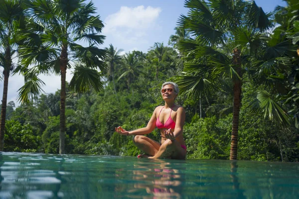 Привлекательная и счастливая азиатка средних лет в бикини, расслабленная в бескрайнем бассейне экзотического курорта в тропических джунглях, сидящая в позе йоги лотоса — стоковое фото