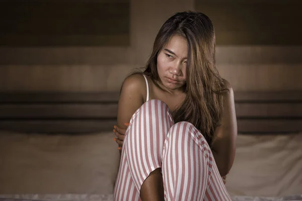 Dramatisches Lifestyleporträt einer jungen, traurigen und depressiven indonesischen Frau im Pyjama, die verzweifelt auf dem Bett sitzt und Schmerzen und Depressionen erleidet, die sich einsam fühlen — Stockfoto