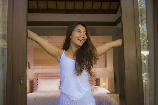 Молодая привлекательная и сексуальная азиатская индонезийская женщина в пижаме просыпается с постели утром, открывая окно на балконе, улыбаясь расслабленно наслаждаясь отпуском — стоковое фото
