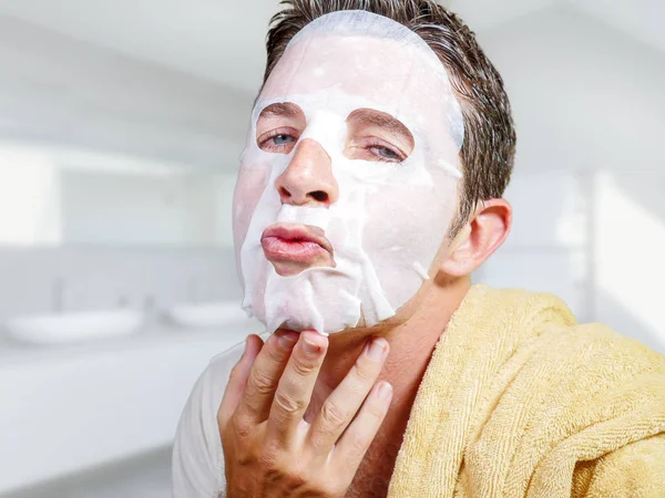 Jong aantrekkelijk en gelukkig gay man in badkamer met behulp van schoonheid papier gezichtsmasker aanraken van zijn gezicht op zoek naar de spiegel glimlachen in mannelijke schoonheid huidverzorging — Stockfoto