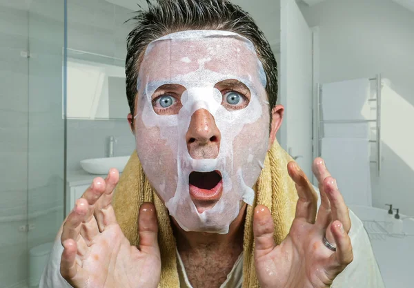 Giovane uomo spaventato e sorpreso a casa utilizzando la pulizia della maschera facciale di carta di bellezza facendo trattamento viso anti invecchiamento guardando se stesso strano e divertente nello specchio del water — Foto Stock