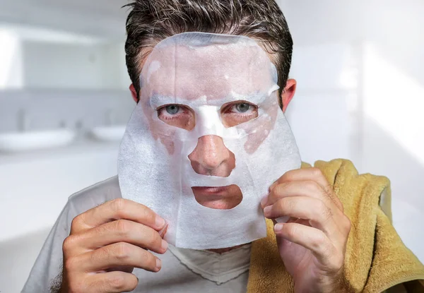 Young scared and surprised man at home using beauty paper facial mask cleansing doing anti aging facial treatment looking himself weird and funny in toilet mirror — Stock Photo, Image