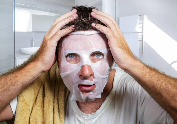 Young scared and surprised man at home using beauty paper facial mask cleansing doing anti aging facial treatment looking himself weird and funny in toilet mirror — Stock Photo, Image