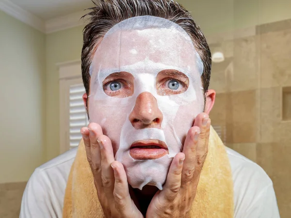Νέος περίεργος και αστείος άνθρωπος στο σπίτι προσπαθεί να χρησιμοποιήσει το χαρτί ομορφιάς προσώπου μάσκα καθαρισμού εκμάθηση αντι θεραπεία γήρανσης σε έκπληκτη έκφραση προσώπου κοιτάζοντας στον καθρέφτη — Φωτογραφία Αρχείου