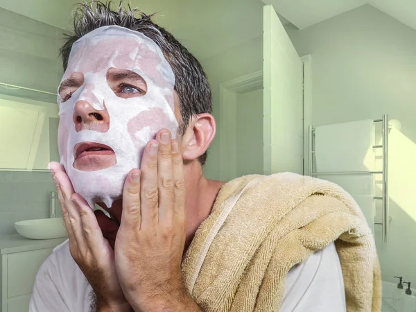 Портрет молодого странного и смешного человека дома, пытающегося использовать косметическую бумажную маску для лица, очищающую обучение антивозрастному лечению в концентрированном выражении лица — стоковое фото