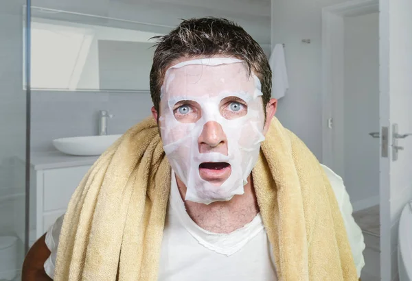 Young funny and messy man applying beauty facial mask looking to the mirror surprised and horrified in skin care aging concept and male cosmetic face treatment — Stock Photo, Image