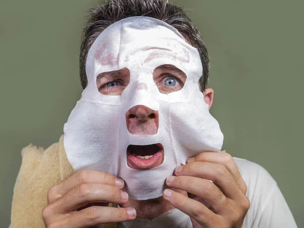 Νεαρός αστείος και βρώμικο άνθρωπος εφαρμογή μάσκα προσώπου ομορφιά κοιτάζοντας στον καθρέφτη έκπληκτος και τρομοκρατημένος στην έννοια φροντίδας του δέρματος γήρανση και ανδρική καλλυντική θεραπεία προσώπου — Φωτογραφία Αρχείου