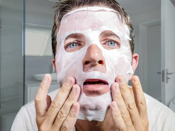 Portrait of young weird and funny man at home trying using beauty paper facial mask cleansing learning anti aging treatment in concentrated face expression — Stock Photo, Image