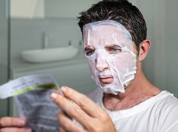 young weird and funny man at home trying using beauty paper facial mask cleansing learning anti aging facial treatment reading the product instructions in bathroom