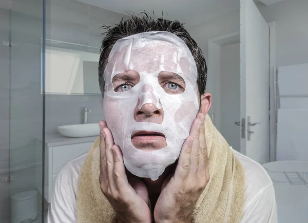 Portrait of young weird and funny man at home trying using beauty paper facial mask cleansing learning anti aging treatment in concentrated face expression — Stock Photo, Image