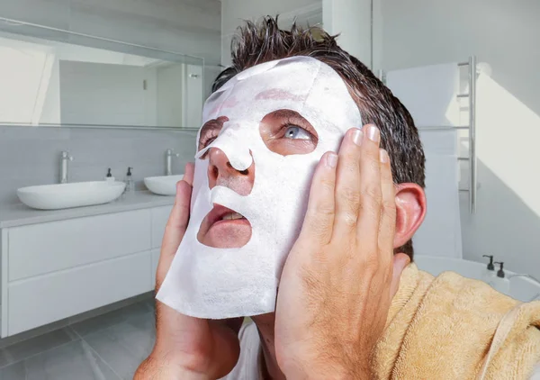 portrait of young weird and funny man at home trying using beauty paper facial mask cleansing learning anti aging treatment in concentrated face expression