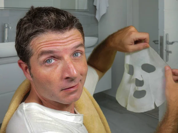 Jeune homme heureux et drôle à la maison en essayant d'utiliser le masque facial en papier beauté nettoyage apprentissage traitement anti-vieillissement dans l'expression du visage bizarre — Photo