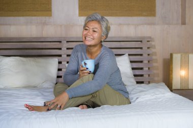 yatakta oturan gri saçlı çekici ve başarılı olgun Asyalı Amerikalı kadının ev portresi kahve içme rahat mutlu ve neşeli gülümseyerek