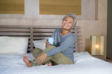 yatakta oturan gri saçlı çekici ve başarılı olgun Asyalı Amerikalı kadının ev portresi kahve içme rahat mutlu ve neşeli gülümseyerek