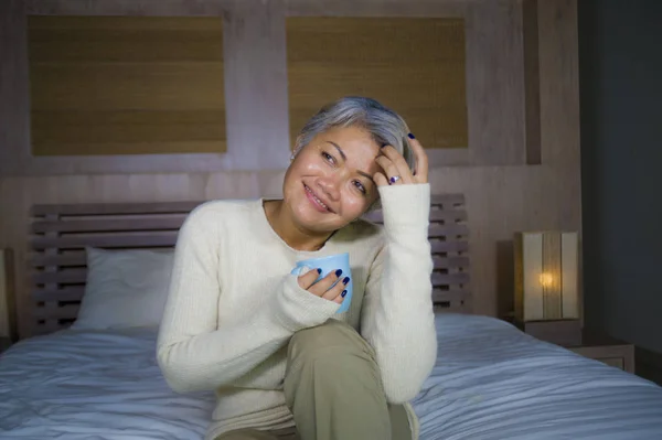 Atractiva y exitosa mujer madura con el pelo gris sentado en la cama bebiendo café relajado sonriendo feliz como mujer de mediana edad disfrutando de estilo de vida doméstico —  Fotos de Stock