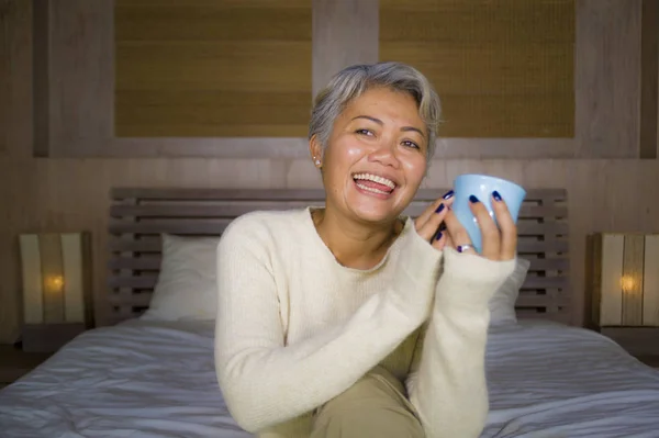 Otthon portréja vonzó és sikeres érett ázsiai-amerikai nő, szürke hajú ül az ágyban iszik kávét nyugodt mosolygó boldog és vidám — Stock Fotó