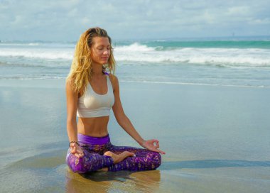 young happy and attractive blond girl doing yoga and concentration exercise outdoors at beautiful beach in relax and meditation practice sitting in lotus position 
