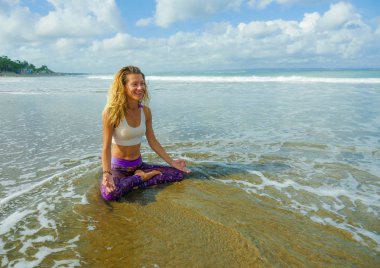 young happy and attractive blond girl doing yoga and concentration exercise outdoors at beautiful beach in relax and meditation practice sitting in lotus position 