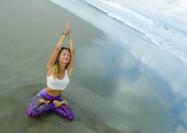 Young attractive and athletic woman sitting in yoga lotus posture and namaste hands position at beautiful beach doing relaxation and meditation exercise in harmony