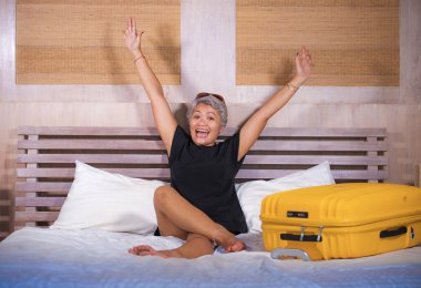 Çekici ve mutlu olgun Asyalı kadın neşeli tatil veya iş gezisi zevk bavul ile yatakta oturan otel odasına gelen gülümseyen