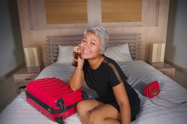 Привлекательная и счастливая зрелая азиатка улыбается веселым прибытием в гостиничный номер, сидя на кровати с чемоданом, наслаждаясь отпуском или деловой поездкой — стоковое фото