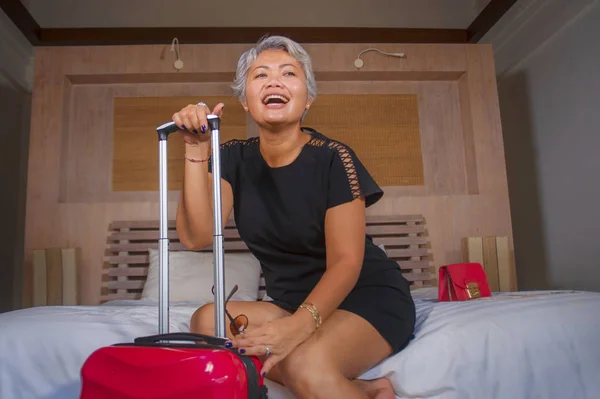 Привлекательная и счастливая зрелая азиатка улыбается веселым прибытием в гостиничный номер, сидя на кровати с чемоданом, наслаждаясь отпуском или деловой поездкой — стоковое фото